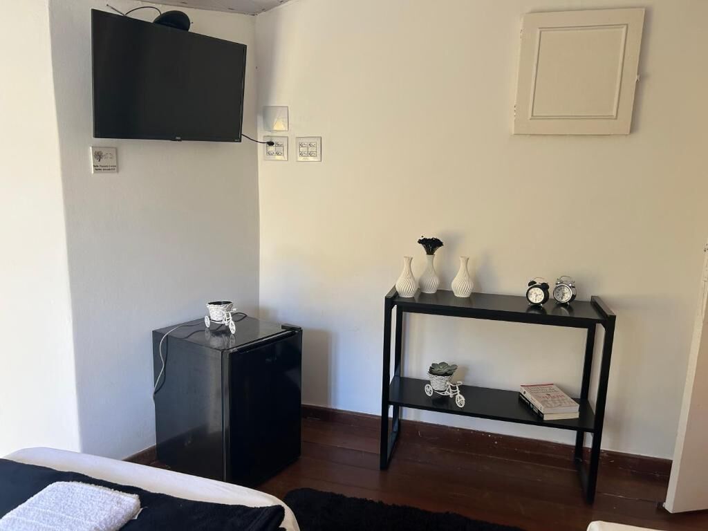 A room with a bed, a black desk, a black TV mounted on the wall, a small table with decorative items, and a white door.