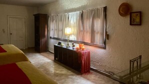 A room with a bed, a bedside table with a lamp, a window with curtains, a hat on the wall, and a door. - Serene Lake Front Beach Cottage in charming Tela (Tela)