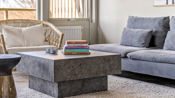 Uma sala de estar moderna com um sofá cinzento, uma mesa de centro com livros, uma poltrona de vime e uma grande janela com persianas.