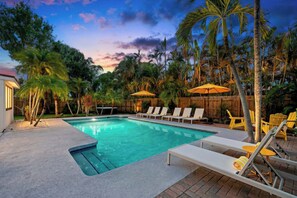 Ein Swimmingpool mit Liegestühlen und Sonnenschirmen, umgeben von Palmen und einem Holzzaun.