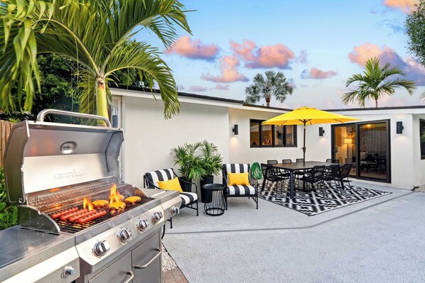 Una parrilla con salchichas y perros calientes, mobiliario exterior con sillas y una mesa, y un paraguas amarillo.