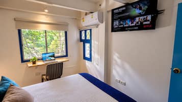 Un dormitorio con cama, escritorio con una computadora portátil, ventana con vistas, aire acondicionado y un televisor de pared que muestra carteles de películas.