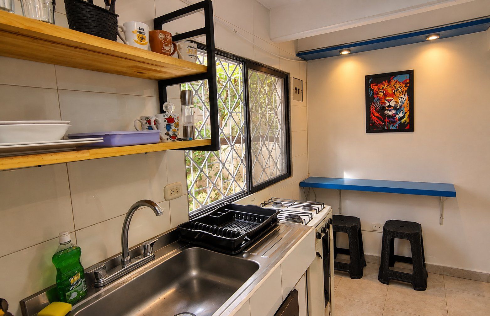 A kitchen with a gas stove, stainless steel sink, and a window with a metal grid. There are stools and a shelf with mugs.
