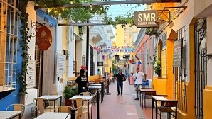Uma rua estreita com mesas e cadeiras, uma placa indicando S.M.R. e edifícios em ambos os lados.