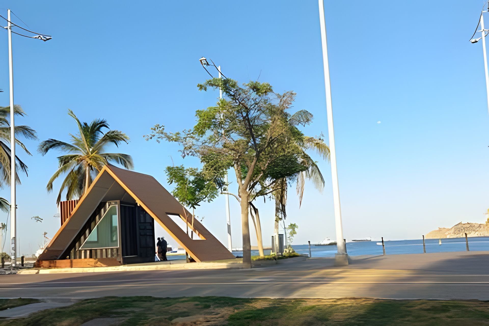 Una estructura arquitectónica moderna con techo triangular, junto a una carretera costera con palmeras y farolas.