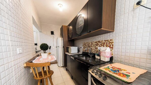 A compact kitchen with white tiled walls, a wooden table, a microwave, and a stove. - Apartamento na Praia Grande a 700m da Praia (Praia Grande)