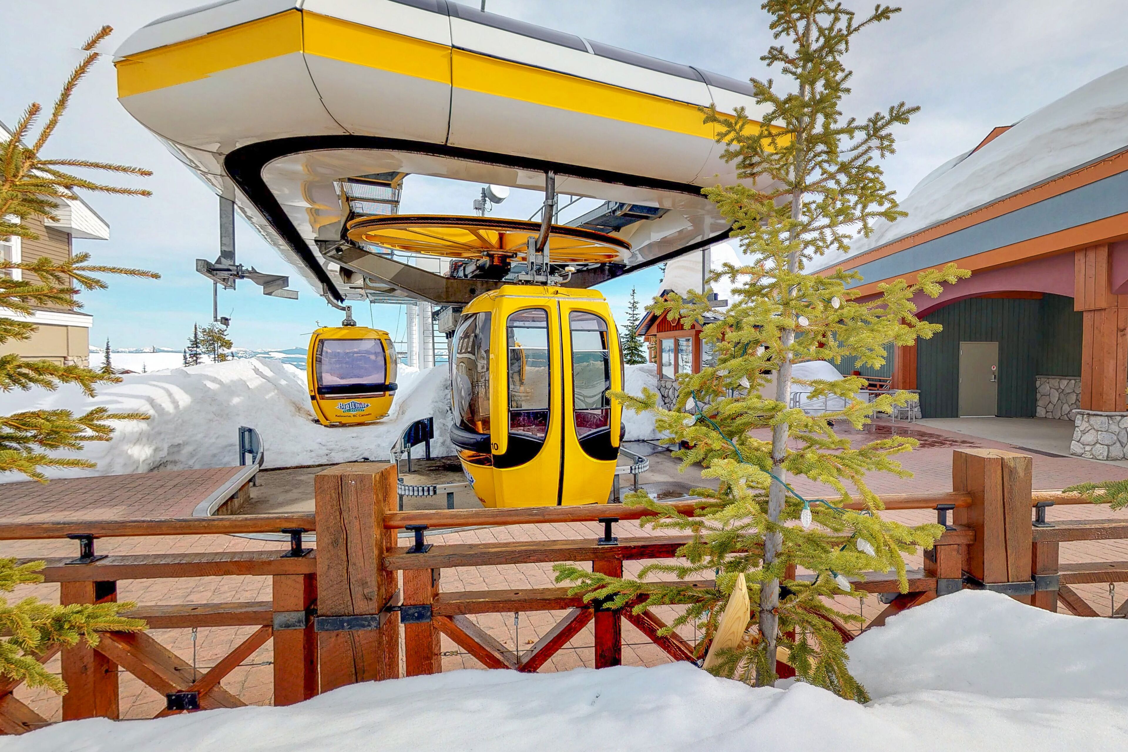 A yellow cable car suspended above a wooden deck with snow on the ground.