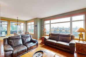 A spacious living room with two leather sofas, a wooden coffee table, and a chandelier. Large windows offer a view of buildings outside. - Glacier Lodge by Okanagan Premier (Beaverdell)