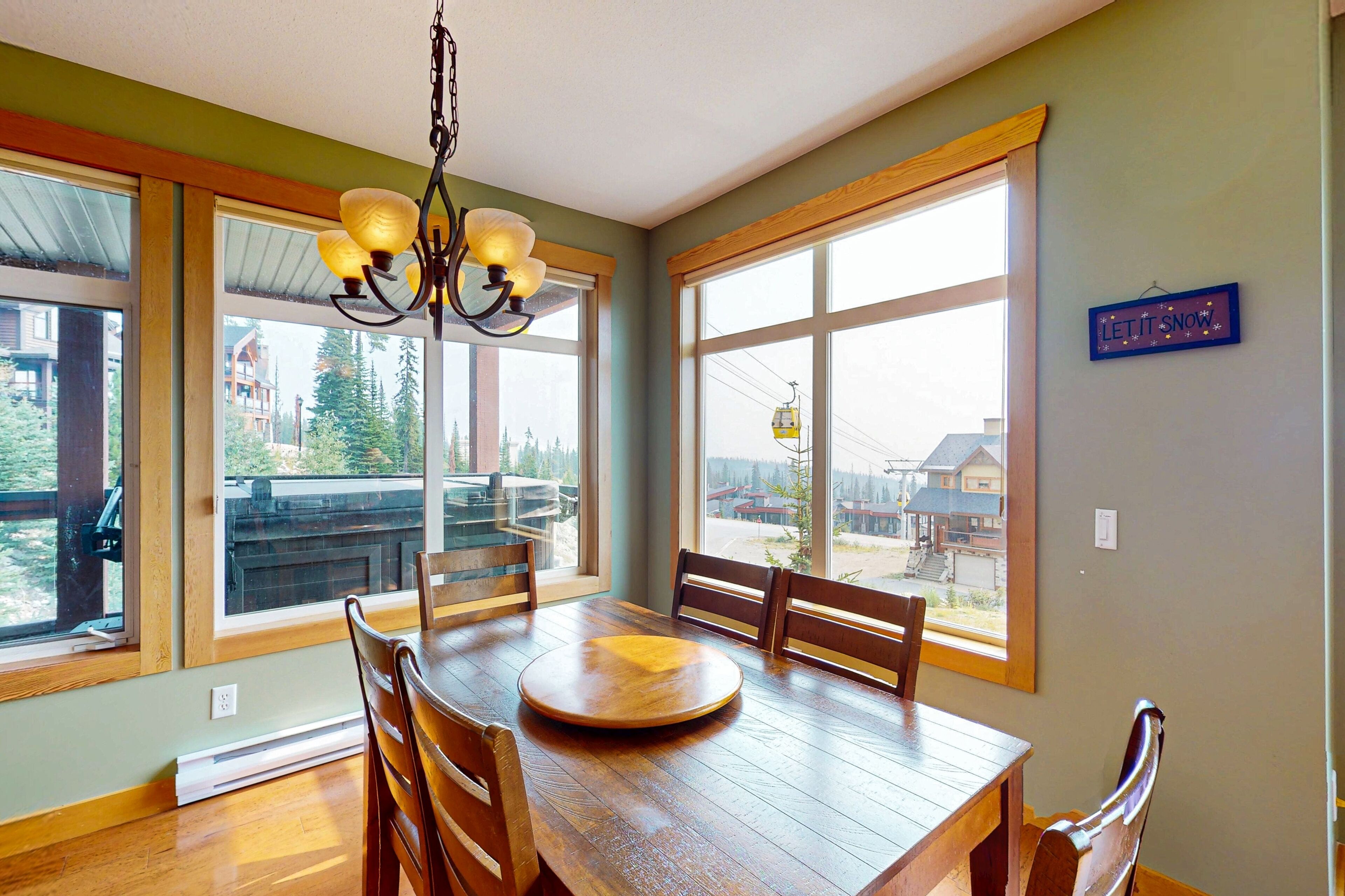 A dining area with a wooden table, chairs, and a chandelier. Large windows provide views of the outside.