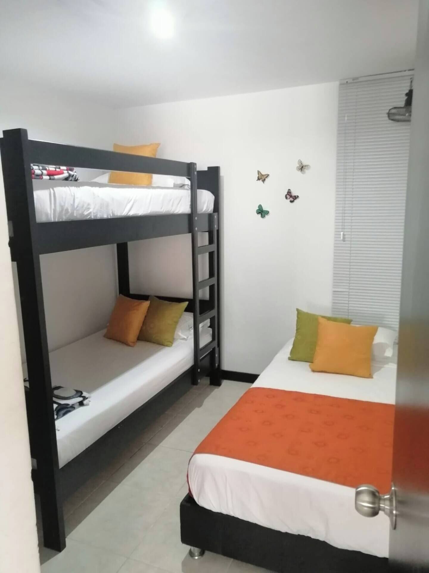 A bunk bed room with a lower bunk and a single bed, both with pillows and a blanket. There are butterfly wall decals and a window with blinds.