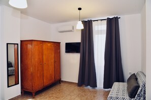 A room with a wooden wardrobe, a flat-screen TV, a sofa with patterned cushions, and a window with dark curtains. - Traditional Cretan house in Limnes – 10 minutes from Agios Nikolaos. (Limne)