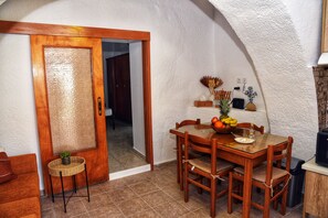 A dining area with a wooden table, chairs, a small side table with a plant, and a door leading to another room. - Traditional Cretan house in Limnes – 10 minutes from Agios Nikolaos. (Limne)