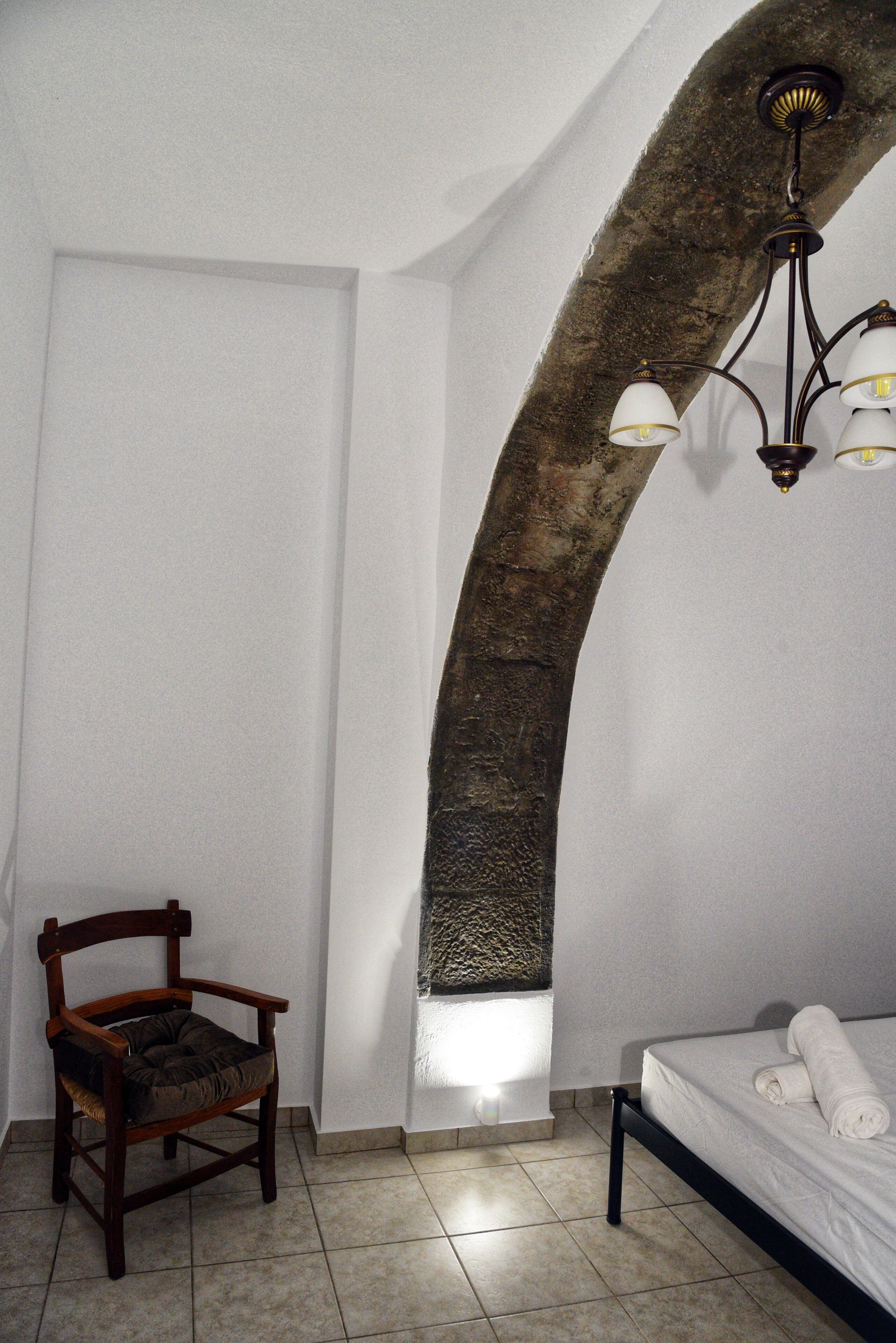A bedroom with a stone arch, a wooden chair, a bed with a rolled towel, and a hanging light fixture.