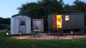 Entorno rústico al aire libre con tres construcciones de madera: una con puerta abierta que muestra un comedor, otra con ventana, y una tercera con puerta cerrada.