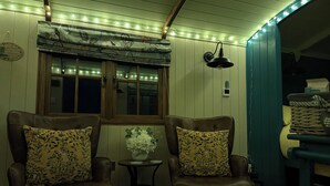 Una habitación con dos sillones de cuero, una mesa pequeña con un florero, una ventana con persianas y una lámpara fijada a la pared.