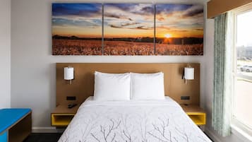 A hotel room with a large bed, a painting of a sunset over a field, two bedside tables with lamps, and a window with curtains.
