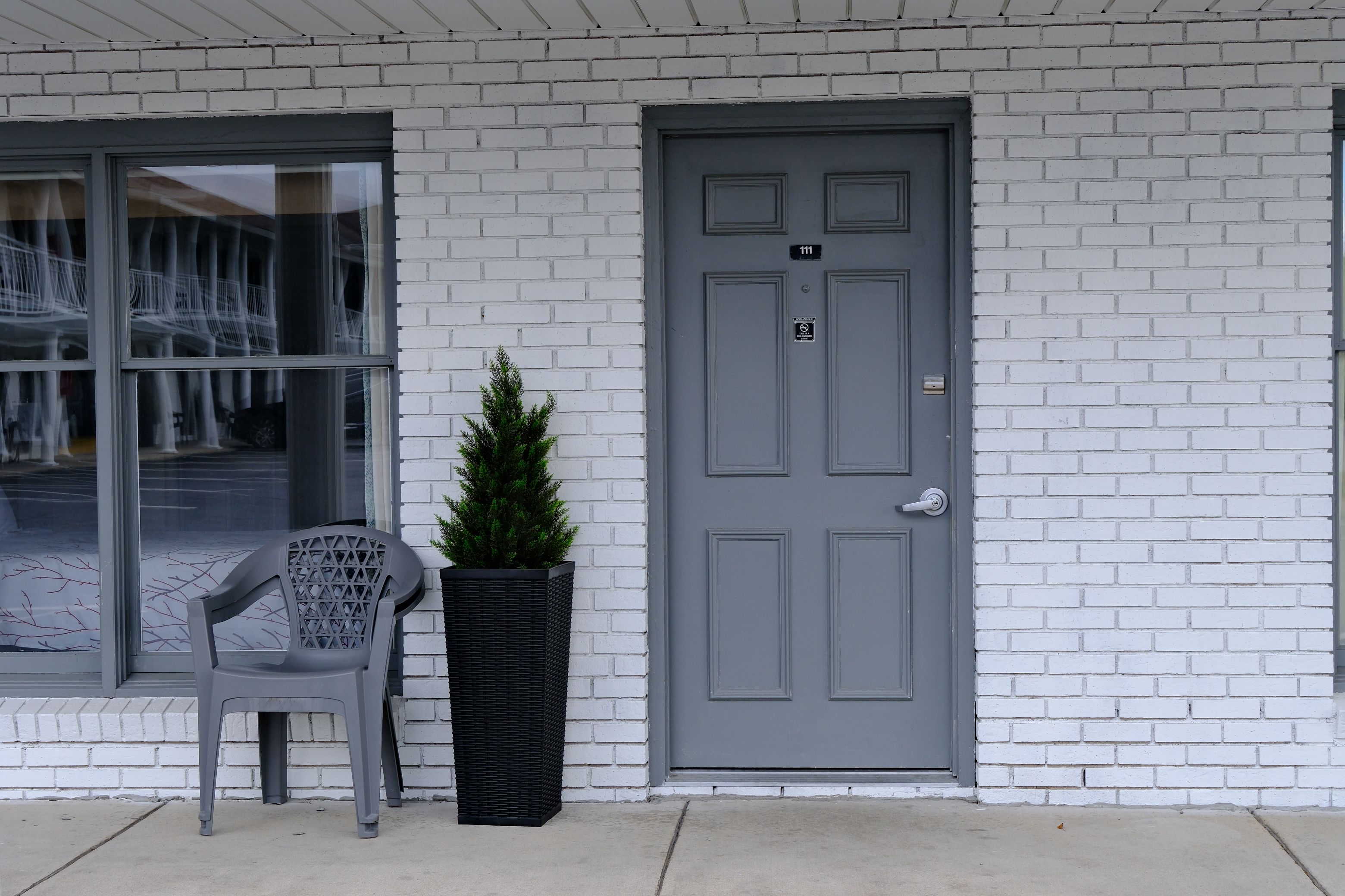 Fachada de edificio con puerta gris, ventana con vista a un estacionamiento y una planta en maceta junto a una silla de plástico.
