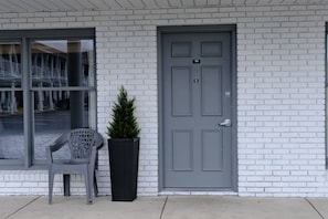 A building facade with a grey door, a window with a view of a parking lot, and a potted plant beside a plastic chair.