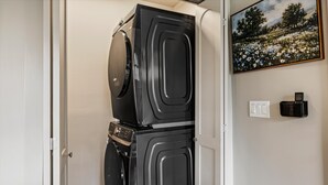 A compact room with a stacked washer and dryer, a power outlet, and a framed landscape painting on the wall. - Historic Hodge House|2BD•2BA|King Beds| Sleeps 6 (Banner Elk)