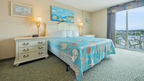 A hotel room with a bed, two bedside tables, a nightstand, a digital clock, a painting, and a view of a marina. - Waterfront Condo | Harbourgate Marina Views (North Myrtle Beach)