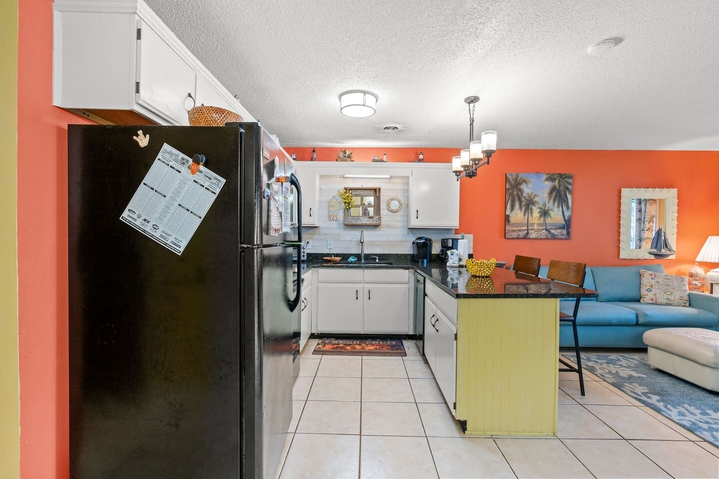 A compact kitchen with white cabinets, a black refrigerator, and a dining area with a blue sofa and yellow table.