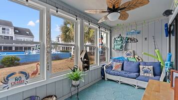 A sunroom with large windows, a ceiling fan, a blue sofa, and a view of a beach and house.