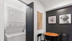 Quarto compacto com máquina de lavar, uma pequena mesa, duas cadeiras e duas fotos emolduradas na parede.