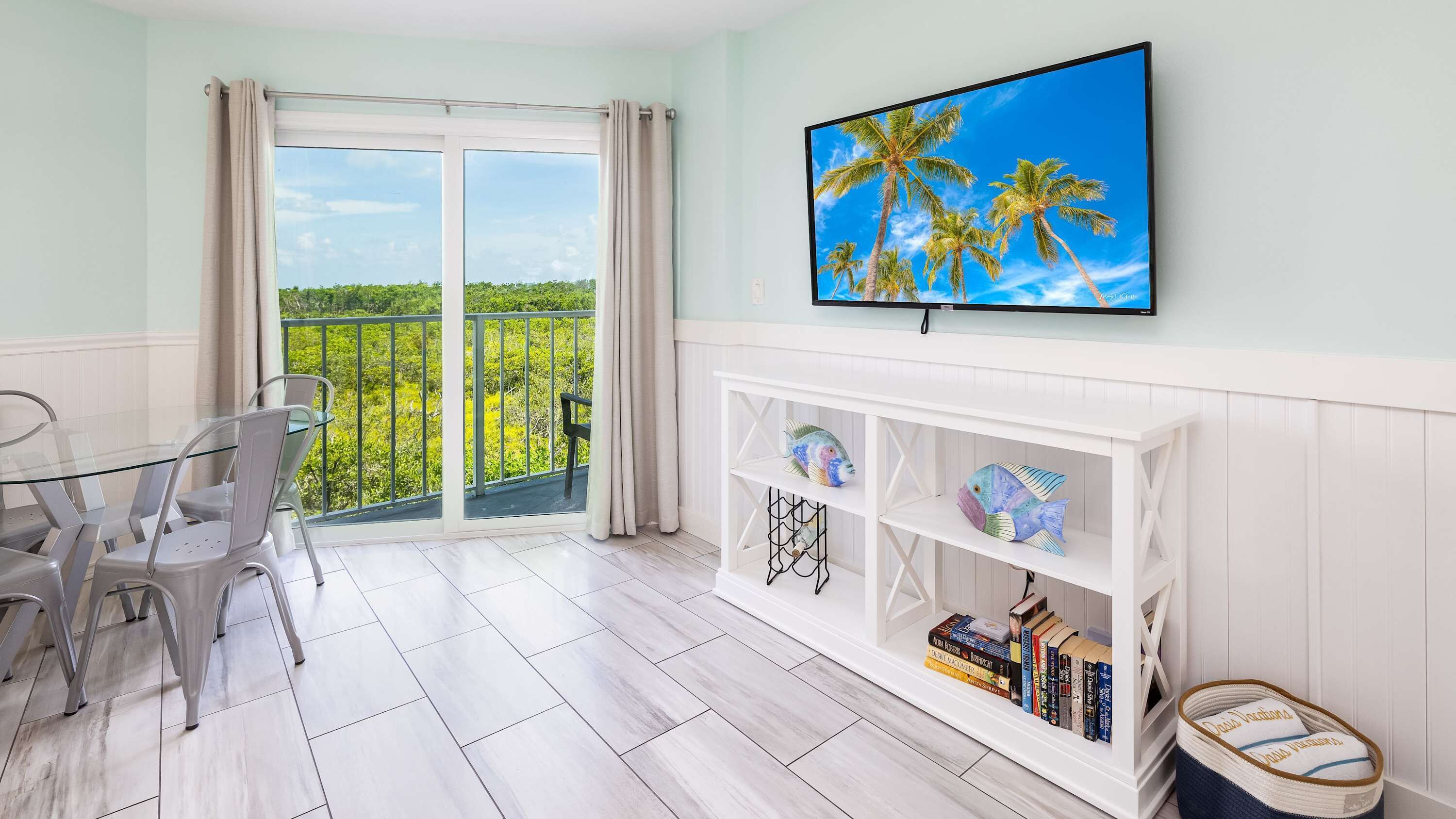 A room with a balcony, a TV mounted on the wall, and a white console table with books and decorative items.