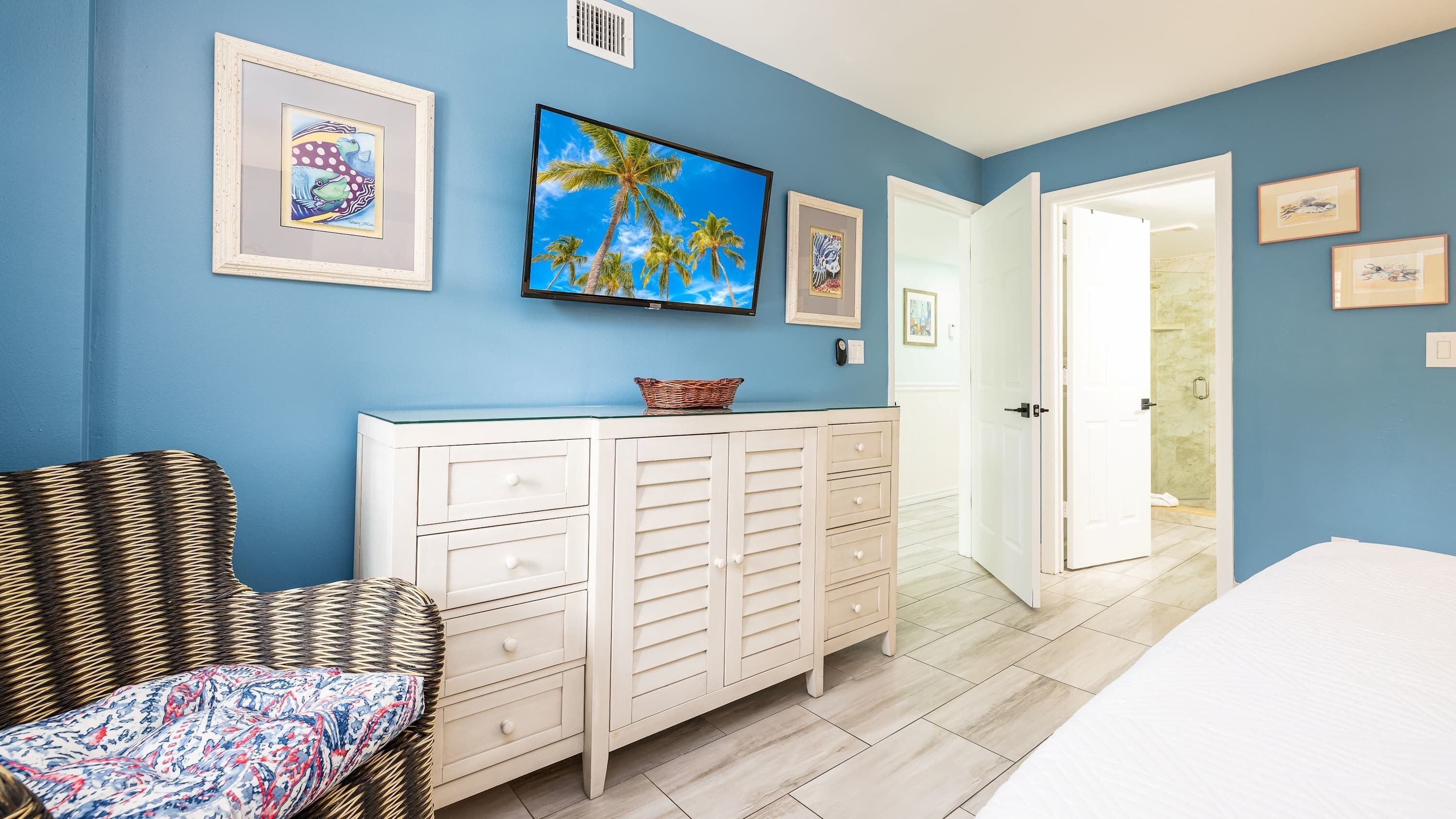 Une chambre à coucher avec un lit, une chaise en osier, une commode blanche et un téléviseur fixé au mur.