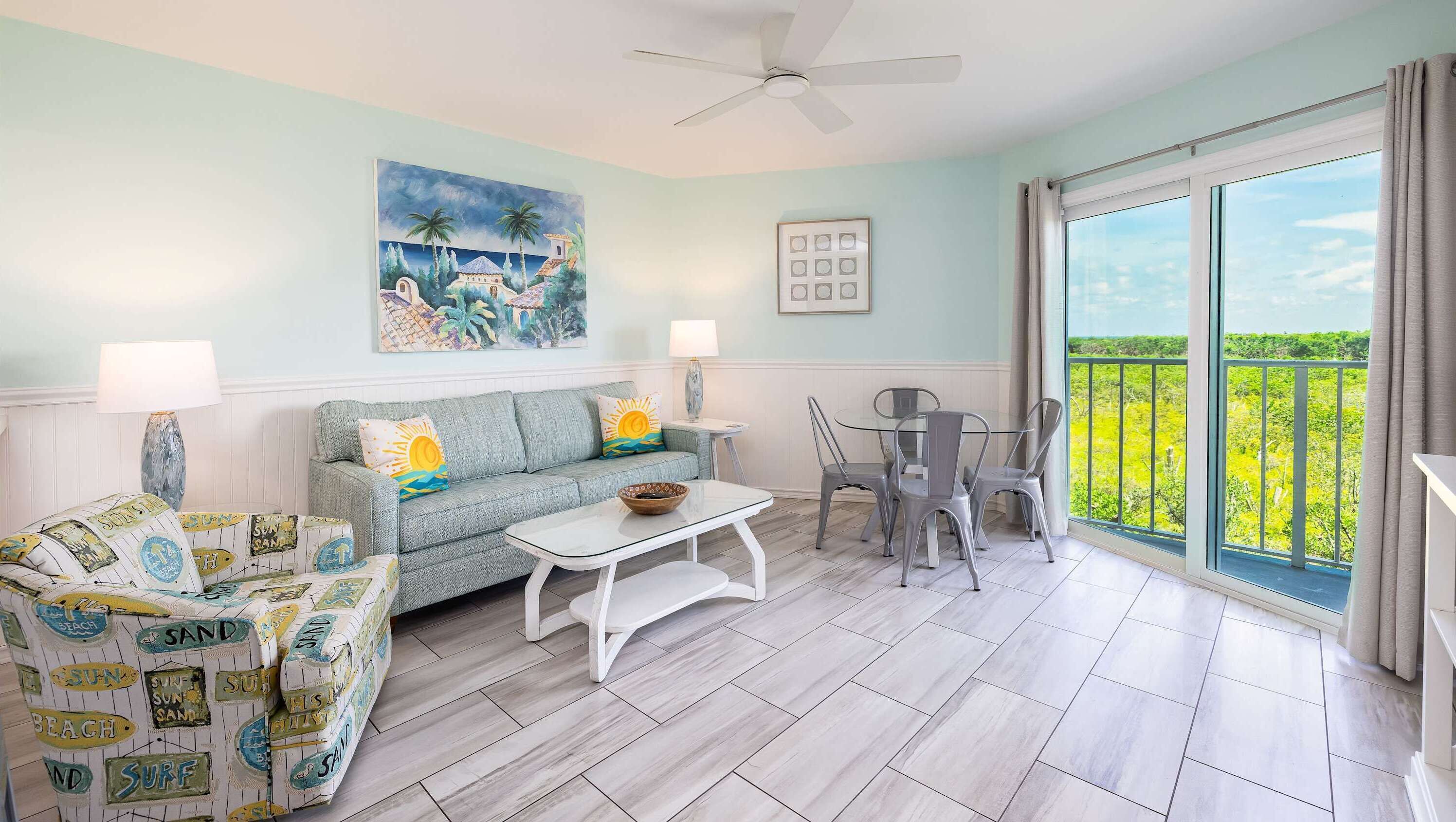 A living room with a sofa, a coffee table, and a dining area with chairs and a table. There is a painting on the wall and a balcony with a view.