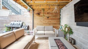 Ein überdachter Außenbereich mit Holzboden, einem Grill, einer Couch, einem Couchtisch und einem Kamin.