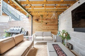 Ein überdachter Außenbereich mit Holzboden, einem Grill, einer Couch, einem Couchtisch und einem Kamin.