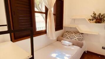 A single bed with a white bedspread, a bedside table, a small rug, and a window with curtains.