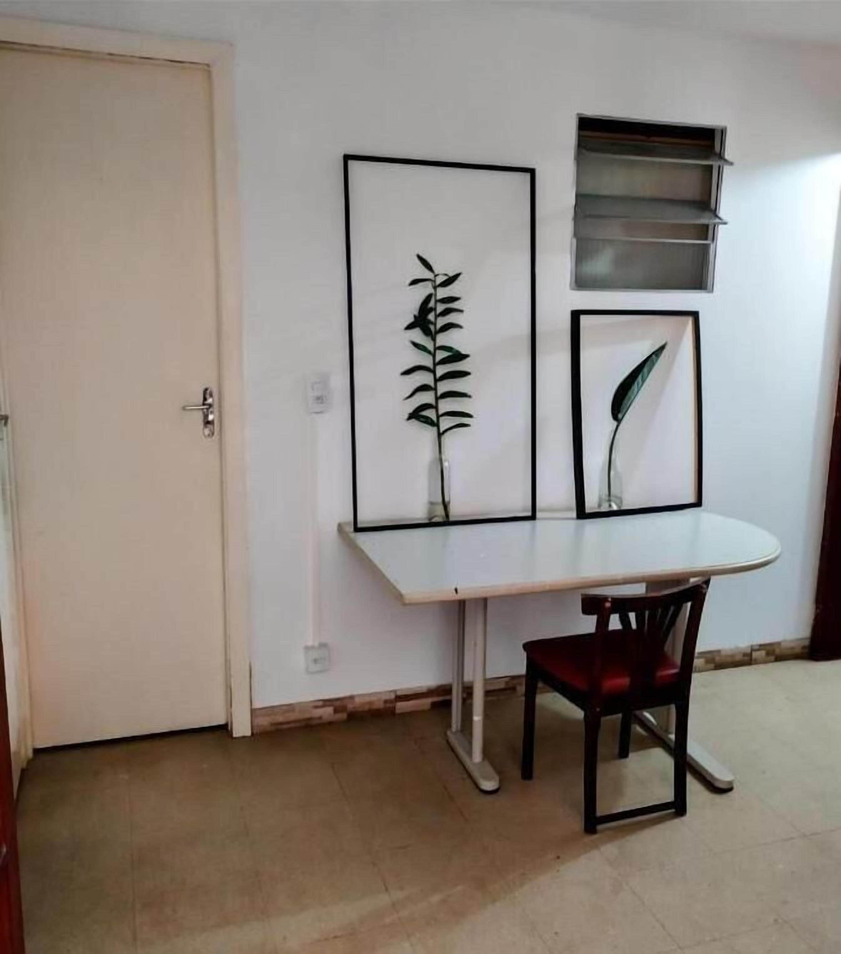 A room with a white door, a white table with a red chair, and two framed botanical prints on the wall.