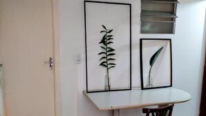 Um quarto com uma porta branca, uma mesa branca e uma cadeira vermelha, além de duas gravuras botânicas emolduradas na parede.