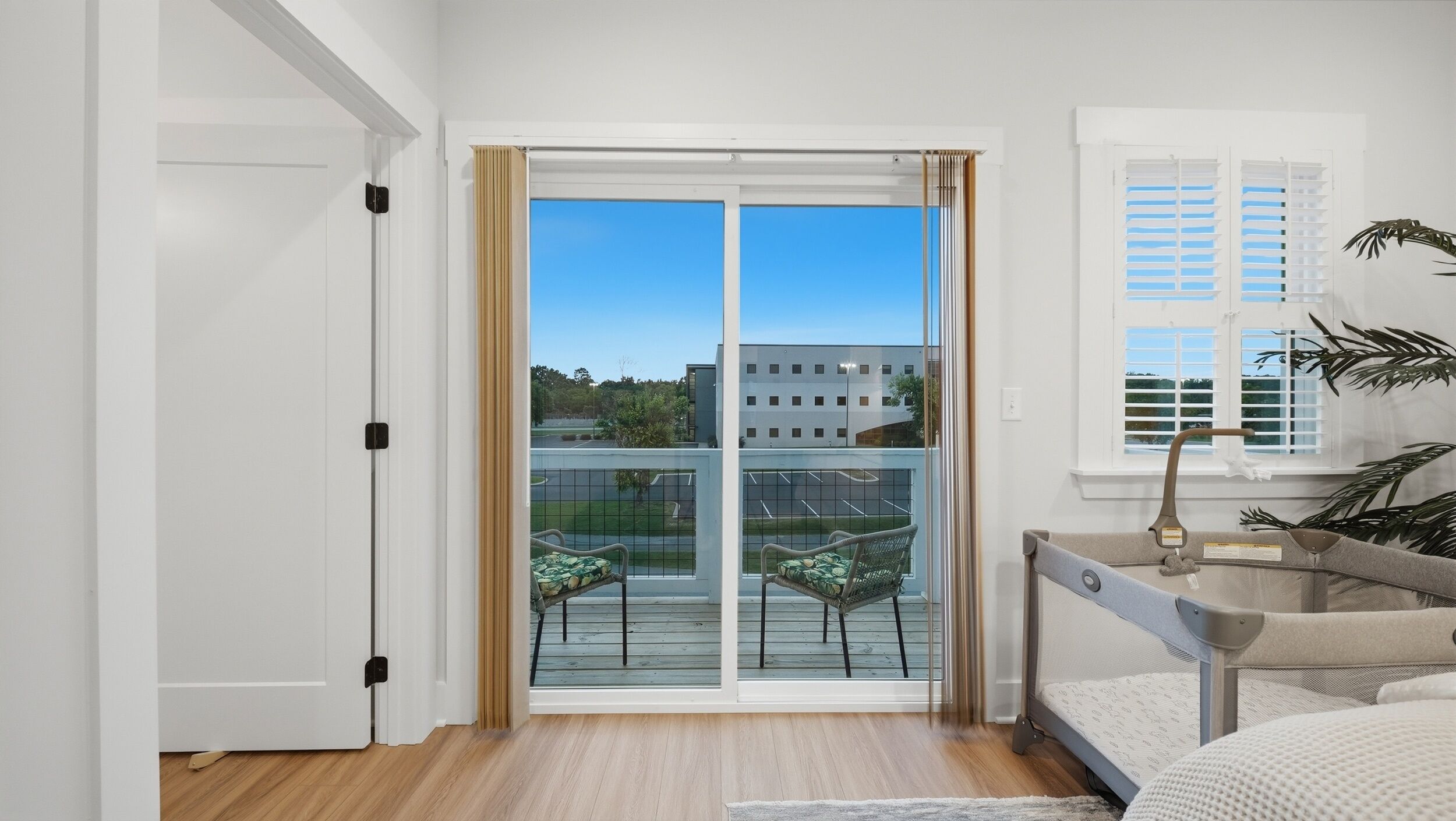 A bedroom with a sliding glass door leading to a balcony, a bed, a small table, and a window with shutters.