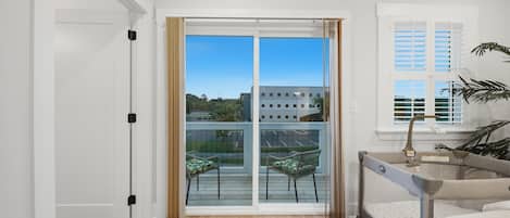 A bedroom with a sliding glass door leading to a balcony, a bed, a small table, and a window with shutters.