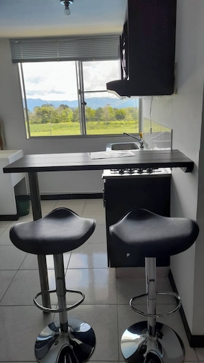 A kitchen with a window view, two bar stools, a black countertop, and a TV mounted on the wall. - Very Affordable Apartment Just 3' from Parque del Café (Montenegro Province)