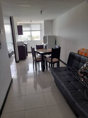 A modern living room with a black leather sofa, a dining area with a table and chairs, and a kitchen area with a refrigerator and microwave. - Very Affordable Apartment Just 3' from Parque del Café (Montenegro Province)