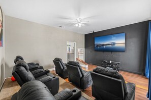 A home theater room with a large flat-screen TV mounted on a dark wall, black leather recliners arranged in a semi-circle, a ceiling fan, and a wooden floor. - 2820 Corey Road (Malabar)