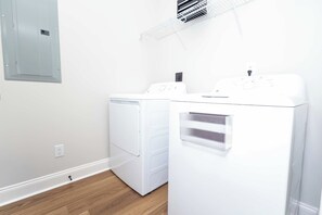 A laundry room with two white washing machines and a dryer, a metal shelf above, and a grey utility cabinet. - The Grove Getaway at Patterson Place (Oxford)