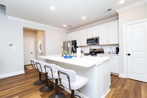 A modern kitchen with white cabinetry, a central island with bar stools, and a microwave. - The Grove Getaway at Patterson Place (Oxford)