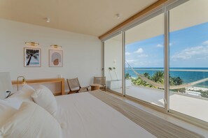 Un dormitorio con cama, dos sillas, un escritorio y un amplio ventanal con vista al océano.