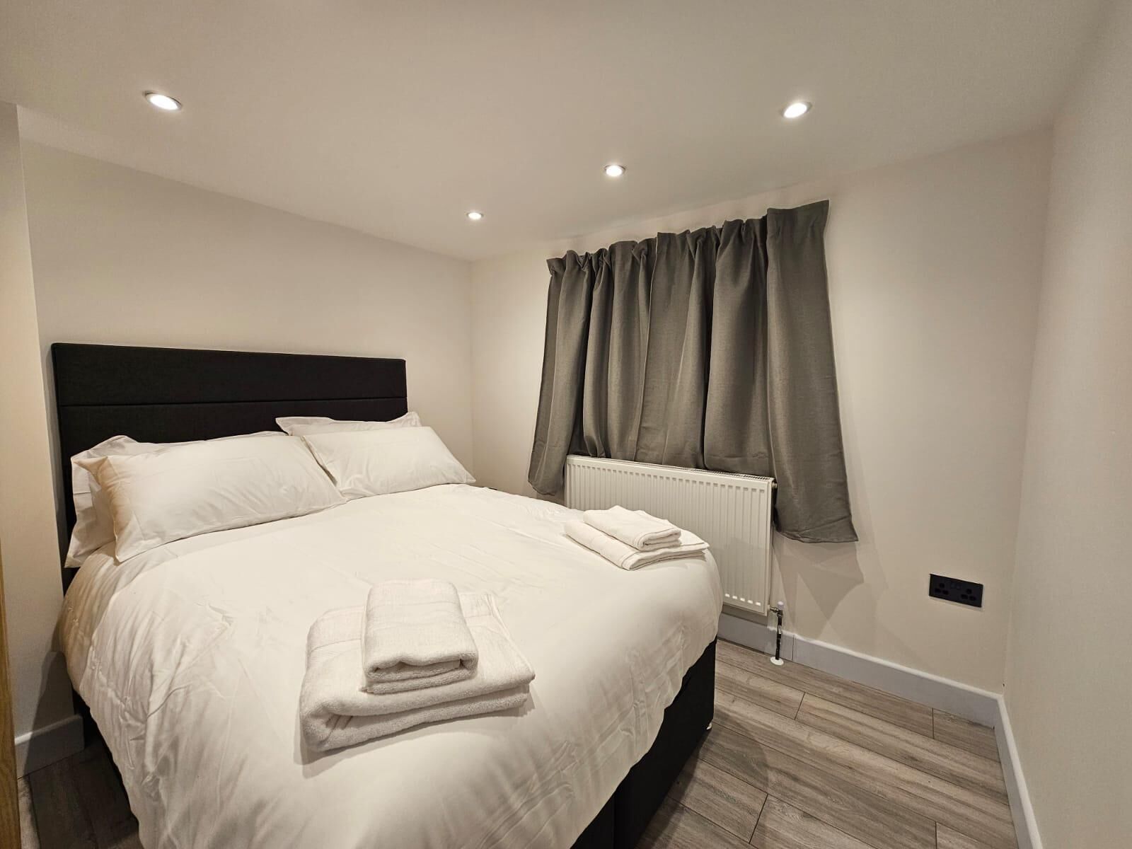 A neatly made bed with pillows and folded towels, a dark headboard, a radiator, and a window with curtains.