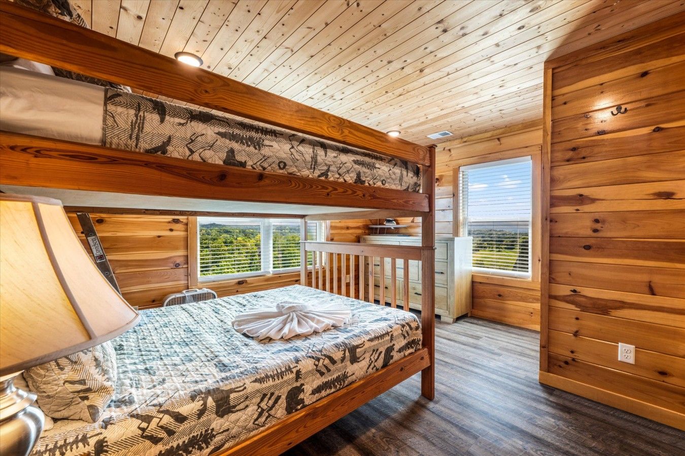 Een stapelbed met een bedsprei met patroon, een bedlampje en een raam met uitzicht op groen.