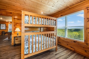 Ein Holzbett mit Etagen, ein Matratzenüberzug mit Bergbildern, ein Fenster mit Blick auf Grünflächen und ein Billardtisch im Zimmer.