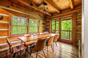A wooden cabin interior with a large dining table, chairs, and a view of the forest through large windows. - 5 Mins to Pigeon Forge! Hot Tub*Game Room*Fire Pit (Pigeon Forge)