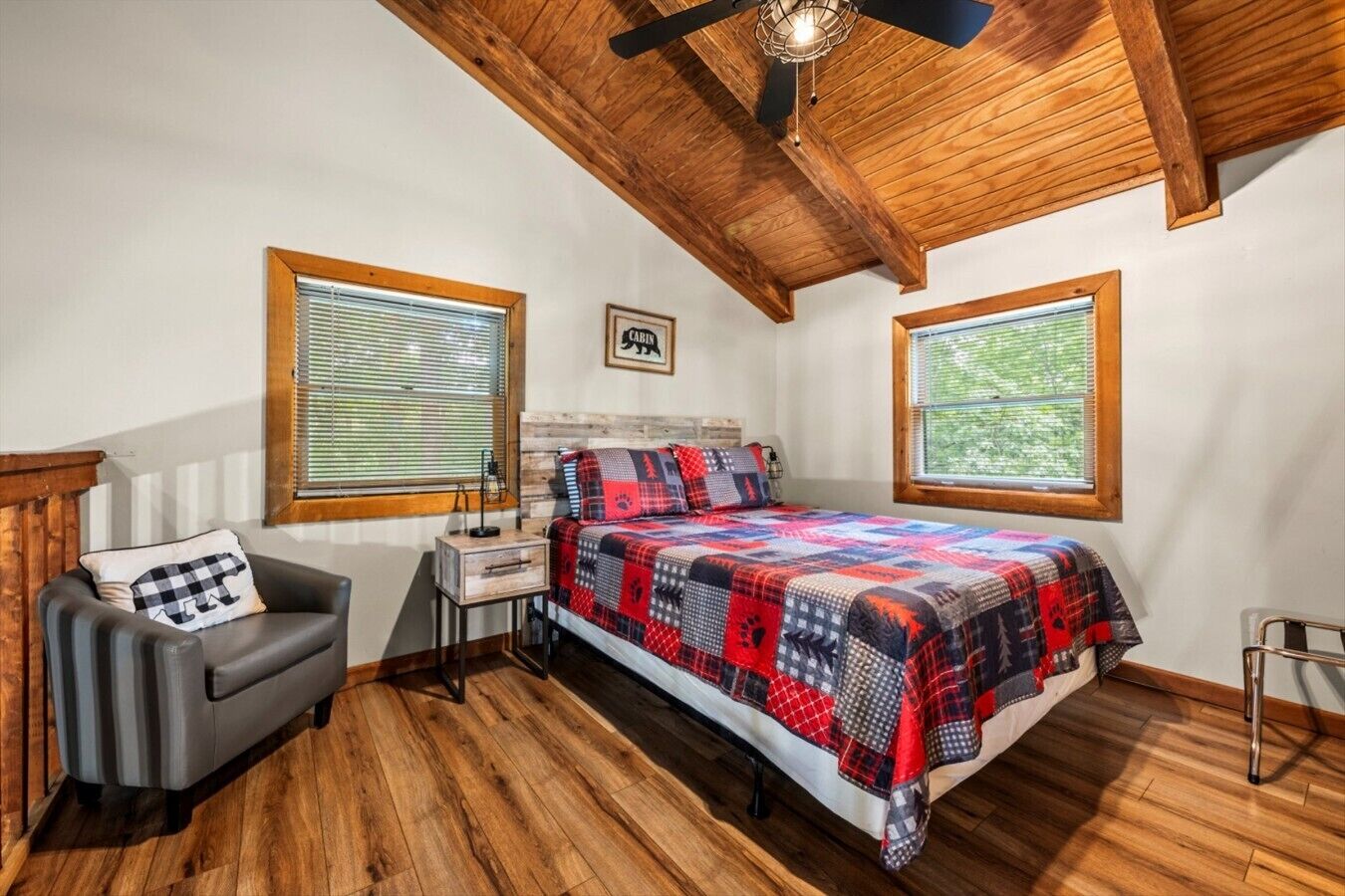 A bedroom with a bed, a chair, a nightstand, a ceiling fan, and two windows with blinds.