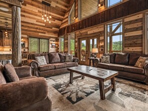 A spacious living room with leather sofas, a coffee table, and a large window providing a view of the outdoors. - 5BR w/Indoor Pool*Theater*Game Room*Hot Tub*Views (Pigeon Forge)