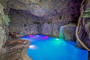 An indoor swimming pool with a rocky cave-like wall, a waterfall feature, and a lounge area with a chair and table. - 5BR w/Indoor Pool*Theater*Game Room*Hot Tub*Views (Pigeon Forge)